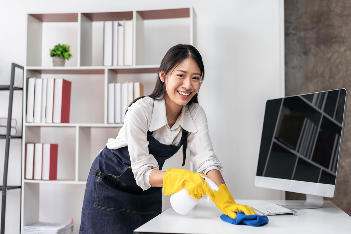 A woman wearing yellow gloves cleans a desk with a spray bottle and cloth in an office, showcasing the thorough work of a professional janitorial service, with bookshelves and a computer in the background.